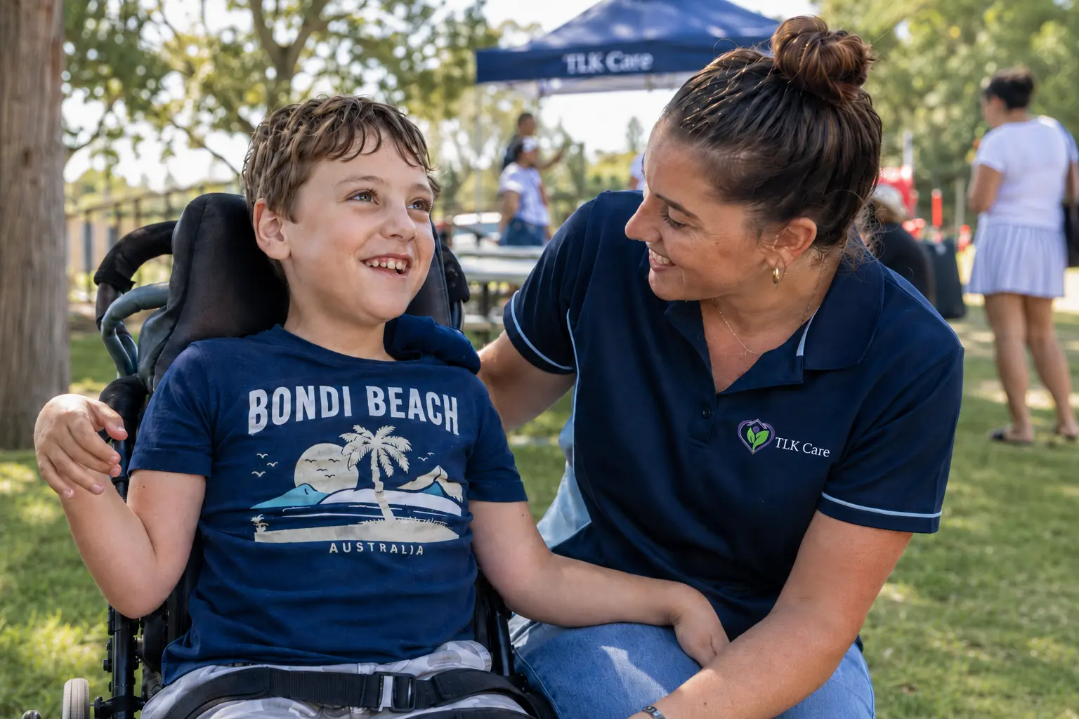 A TLK Care support worker smiling warmly with a young wheelchair user at an outdoor NDIS community event in Sydney.